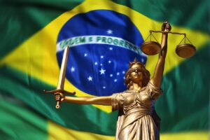 Símbolo da justiça (mulher de olhos vendados segurando uma espada e uma balança) com a bandeira brasileira ao fundo.