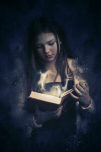 Imagem em tons escuros de uma mulher olhando para um livro aberto em suas mãos. De dentro do livro sai uma espécie de fumaça luminosa.